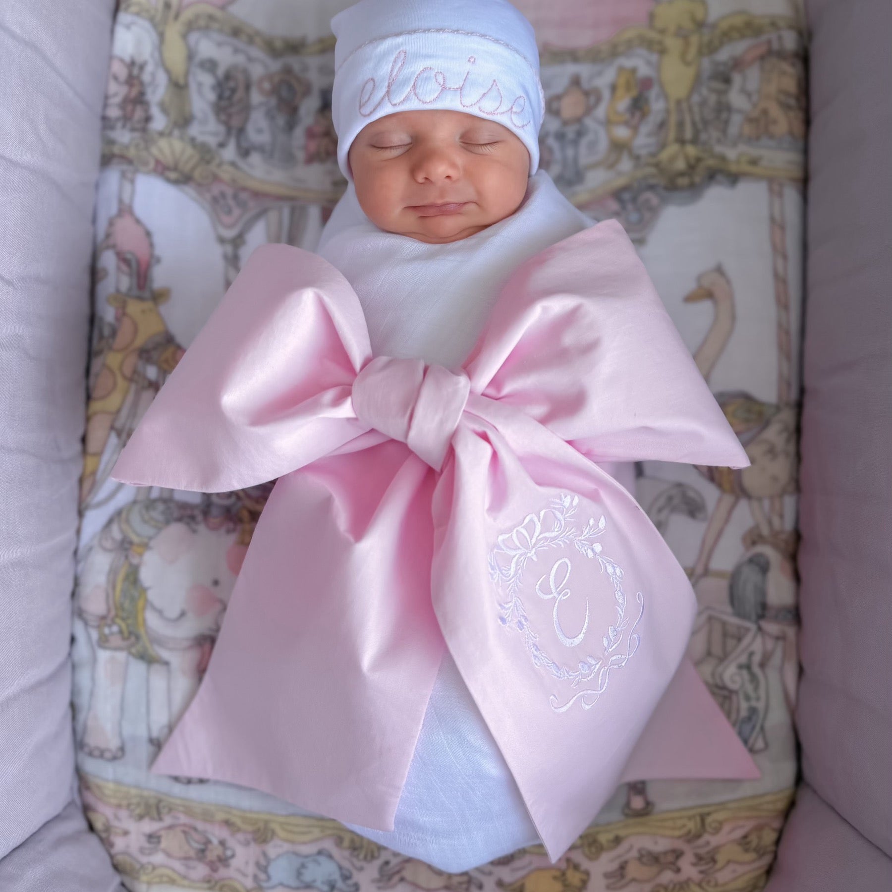 Sleeping baby wrapped in a pink bow blanket with an Eloise embroidered hat